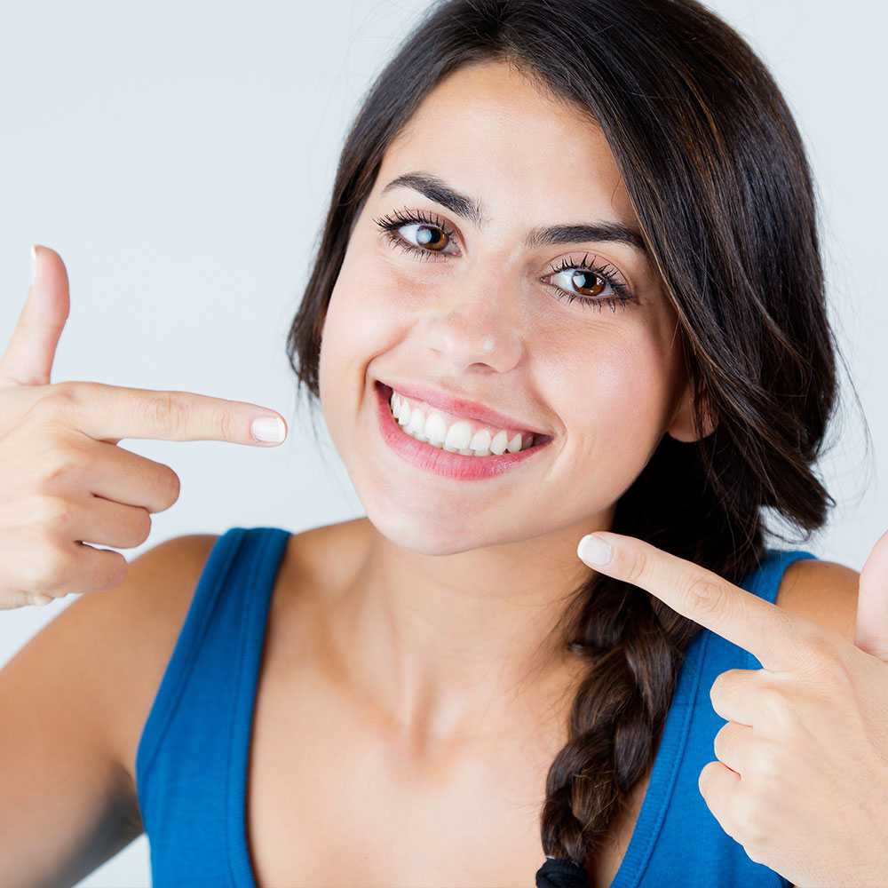 A young woman with a radiant smile pointing to her teeth, showcasing them against a white background.