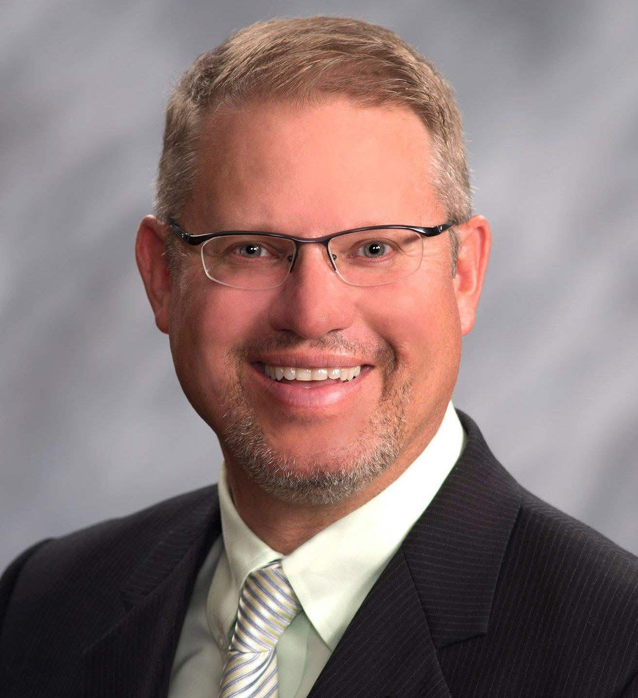 The image shows a man wearing glasses, smiling, with short brown hair, dressed in a suit and tie, posing for a portrait-style photograph.